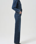 A person stands in profile against a plain light background wearing the Harper Straight Jean and a dark blue denim jacket, both crafted from organic cotton.