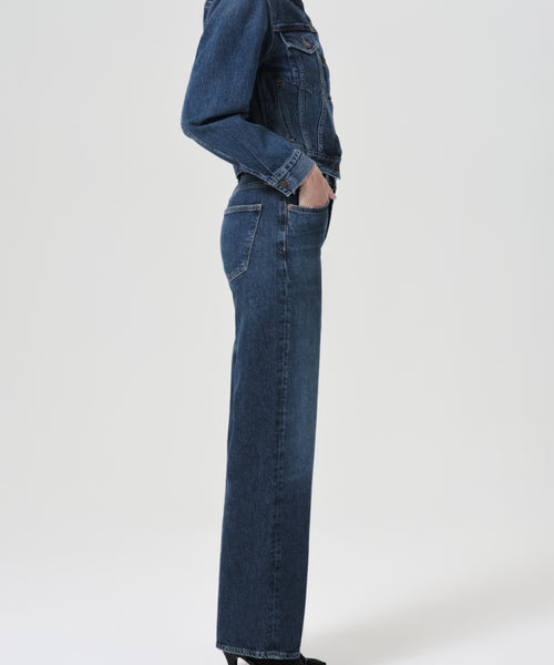A person stands in profile against a plain light background wearing the Harper Straight Jean and a dark blue denim jacket, both crafted from organic cotton.