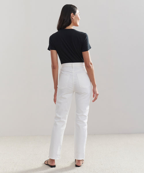 A woman with shoulder-length dark hair faces away, wearing a black short-sleeve t-shirt, High Rise Stovepipe Jean in vintage denim, and black sandals. She stands on a light-colored floor against a plain background.