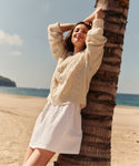 Woman in a beige sweater and white skirt leaning against a palm tree on a beach.