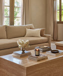 A cozy living room with a beige sofa, the Alpaca Basketweave Lumbar Pillow, and a throw blanket. A travertine coffee table holds stacked books, a candle, a small vase of white flowers, and a ceramic dish. Large windows fill the space with natural light.