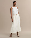 A woman stands against a beige background wearing a white sleeveless top, the cream Annabel Skirt with a vintage-inspired high waist, and white sandals. Her hair is pulled back and she poses with one hand at her side.