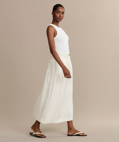 A woman wearing a sleeveless white top, the cream Annabel Skirt, and black flip-flops stands sideways against a plain beige background.