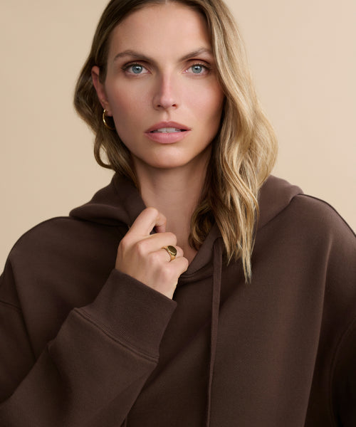 A woman with wavy blonde hair, wearing the Bay Hoodie made from brushed fleece and gold hoop earrings, looks slightly to the side while holding the hoodie's collar. The background is beige.