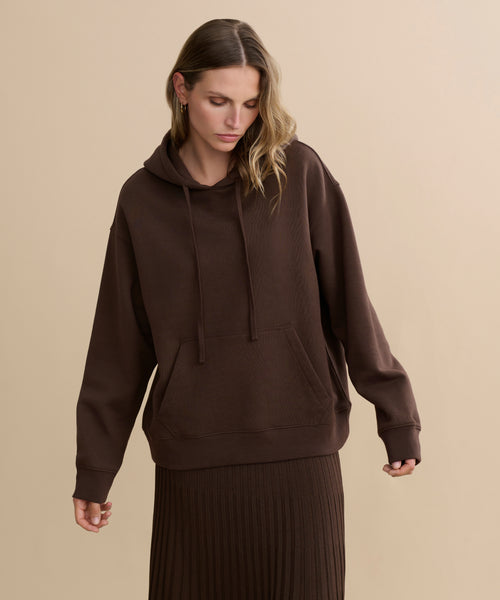A woman wears the Bay Hoodie, a loose-fitting chocolate brushed fleece top, paired with a matching ribbed skirt. She stands against a plain beige background, looking down with her hands slightly out to the sides.