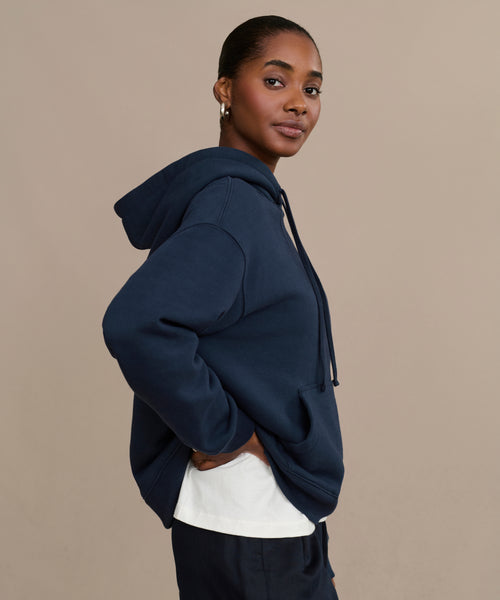 A woman with her hair pulled back poses against a beige background, looking at the camera. She wears a navy Bay Hoodie over a white shirt.