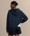A person with short hair in a bun is seen from behind, wearing the Bay Hoodie in navy brushed fleece with matching shorts and one hand on their hip, set against a plain beige background.