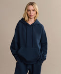 A blonde woman wears a loose-fitting navy Bay Hoodie with matching pants, standing with hands in the pocket against a plain beige background.