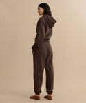 A person stands sideways, facing left, wearing a matching dark brown brushed fleece hoodie and Bay Sweatpant with hands in pockets, plus brown slippers, against a beige background.