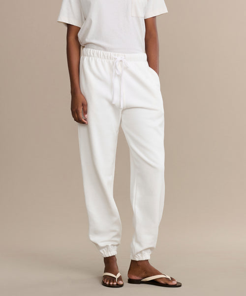 A person wearing a white t-shirt and Bay Sweatpant joggers, crafted from brushed fleece with an elastic waistband and drawstring, stands in sandals against a neutral background. The persons head is not shown.