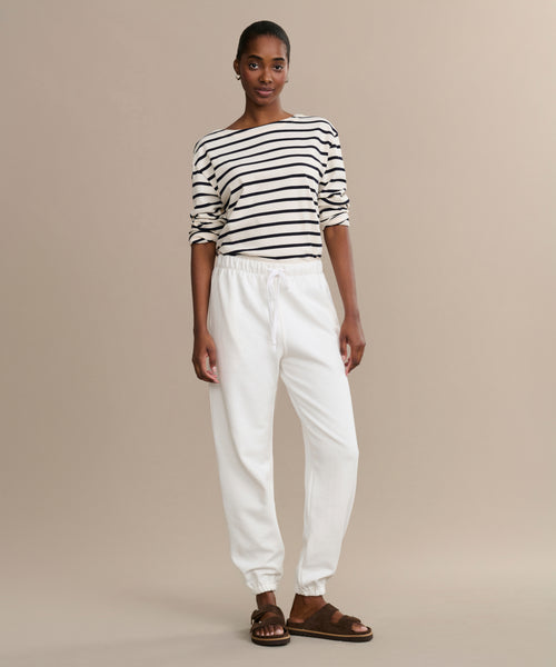 A woman stands against a beige background, wearing a black and white striped long-sleeve top, Bay Sweatpant in white, and brown sandals. She looks confidently at the camera with her hands relaxed at her sides.
