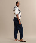 A woman stands sideways wearing a white shirt, navy Bay Sweatpant joggers, sandals, and a cream sweater with navy stripes draped over her shoulders. The background is plain beige.