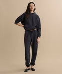 A woman with long dark hair poses casually against a beige background, wearing a black hoodie and the Bay Sweatpant in athletic brushed fleece, paired with black flats.