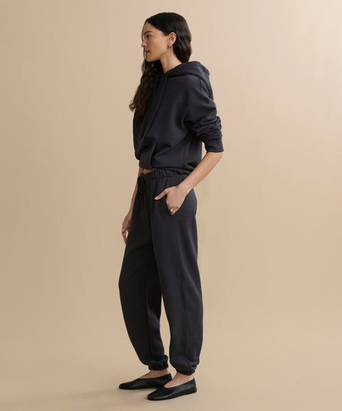 A woman stands in a relaxed pose with her hands in her pockets, wearing a loose dark hoodie and Bay Sweatpant athletic sweats made from brushed fleece, paired with black flats. She has long, wavy hair and is set against a plain beige background.