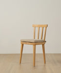 The Hampton Chair Cushion, upholstered in beige Belgian linen with a removable cover, rests on a simple wooden chair placed on a light wood floor against an off-white wall.