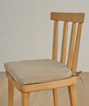 A wooden chair with a beige Hampton Chair Cushion made of Belgian linen, featuring a removable cover tied to the seat, sits on a wooden floor against a plain light wall.
