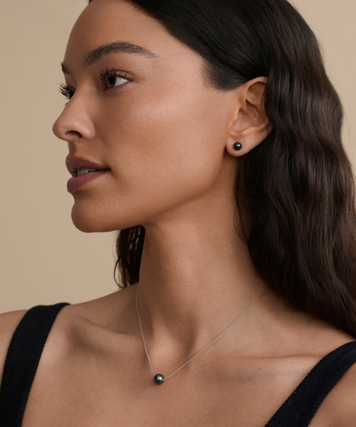 Woman with long, wavy dark hair wears a black top, black pearl stud earring, and the Philo Pearl Necklace—a delicate gold necklace with a black pearl pendant—looking to the left against a beige background.