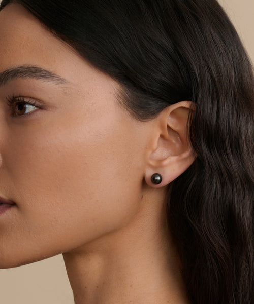 Close-up of a woman's face and ear highlights her smooth skin, dark wavy hair, and Medium Loren Pearl Studs against a neutral background.