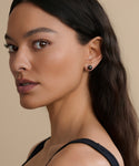 A woman with long, wavy dark hair poses in a black top, wearing Medium Loren Pearl Studs from Modern Elegance Jewelry. She faces the camera at a slight angle against a neutral beige background.
