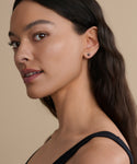 A woman with long dark hair and brown eyes wears a black sleeveless top and chic Jenni Kayne Jewelry Small Loren Pearl Studs, looking over her shoulder against a beige background.
