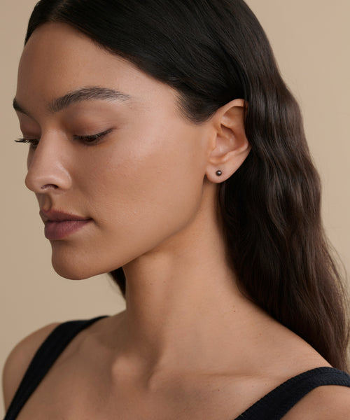 A woman with long, wavy dark hair looks downward in profile, wearing a black sleeveless top and the Small Loren Pearl Studs. The background is beige with soft lighting.