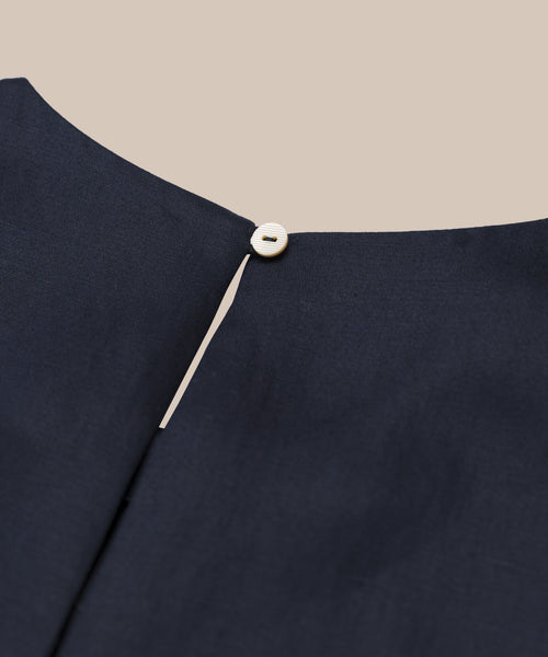 Close-up of the back neckline of the Blair Dress in dark navy, showcasing a subtle linen texture, small keyhole opening, and round cream button fastening. Perfect for spring. Background is solid light beige.