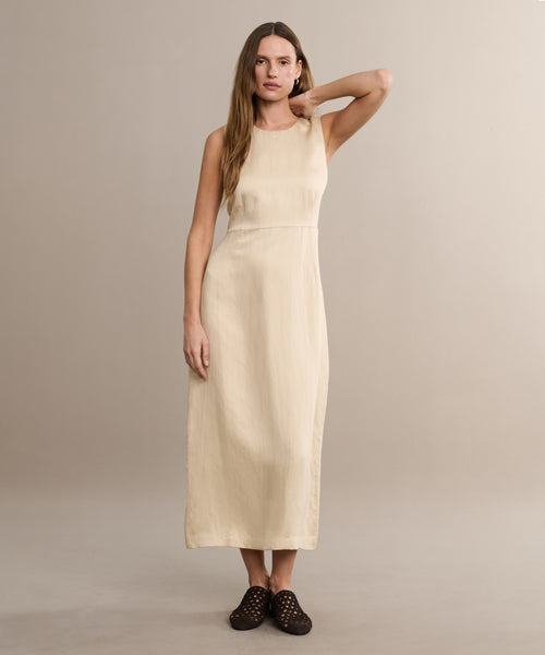 A woman with long hair stands against a beige background wearing the sleeveless, ankle-length Blair Dress—a cream linen dress—plus black woven slip-ons. One arm is bent with her hand touching her head.