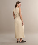 A woman faces away, wearing the sleeveless, ankle-length Blair Dress in beige linen—ideal for spring—paired with black flats featuring cut-out details, set against a plain light background.