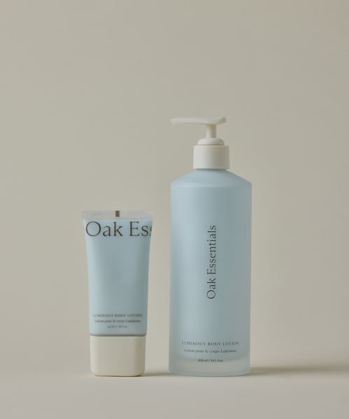 The Luminous Home and Away Duo features two light blue Oak Essentials Luminous Body Lotion bottles—one with a flip cap and one with a pump—perfect for silky-soft skin at home or on the go, set against a plain beige background.