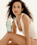 A woman with wavy dark hair in a white sleeveless top sits holding the Luminous Home and Away Duo, a travel-friendly set against a neutral background—ideal for achieving hydrated, silky-soft skin anywhere.