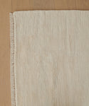 A close-up of the Bowery Rug, a beige handwoven piece with fringed edges, highlighting its subtle pattern and soft texture on a light wooden floor.