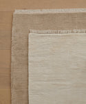 Two rectangular Bowery Rugs, one light beige and one brown, are layered on a light wood floor. The smaller beige Bowery Rug is placed diagonally over the larger brown Bowery Rug, both featuring fringed edges.