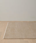 The Bowery Rug, a large rectangular handwoven beige rug, sits on a light wooden floor against an off-white wall in a minimalistic room.