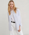 A woman modeling a light blue long-sleeve button-up shirt over a white tee, paired with white trousers and a black belt, against a plain light gray studio background.