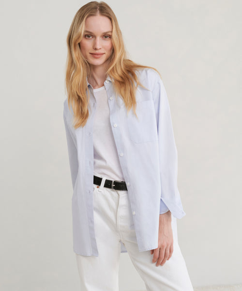 A woman modeling a light blue long-sleeve button-up shirt over a white tee, paired with white trousers and a black belt, against a plain light gray studio background.