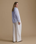 A woman stands in profile wearing a light blue long-sleeve blouse with wide-leg off-white trousers, photographed against a solid warm beige studio background.