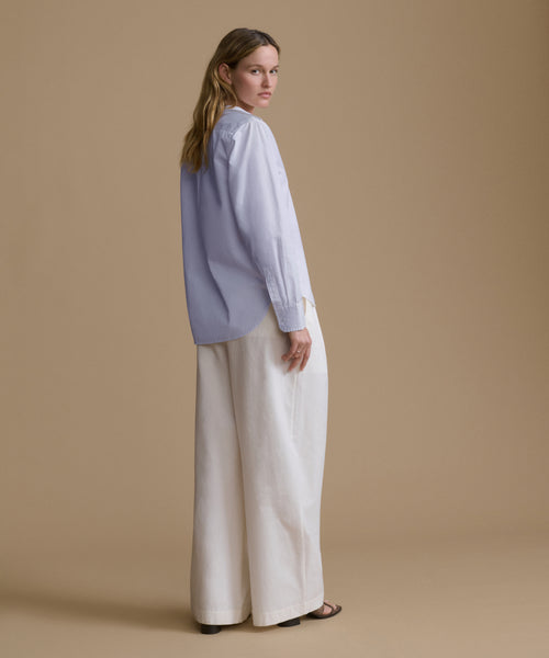 A woman stands in profile wearing a light blue long-sleeve blouse with wide-leg off-white trousers, photographed against a solid warm beige studio background.