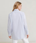 A woman shown from the back wearing a light blue long-sleeve button-up shirt and white pants against a plain light beige studio background.