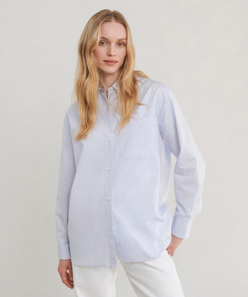 A woman modeling a light blue long-sleeve button-up shirt with white pants, photographed against a plain light gray studio background.