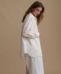 Woman wearing a stripe button up shirt with white pants against a beige background.