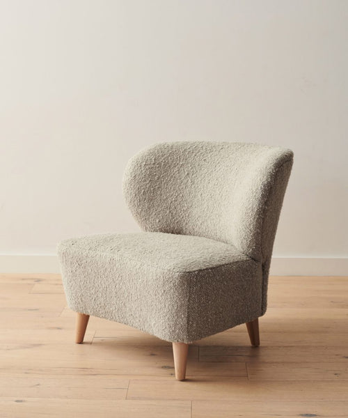 The Brentwood Chair features a modern armless design with light beige Belgian wool upholstery and four wooden legs, shown on a light wood floor against an off-white wall.