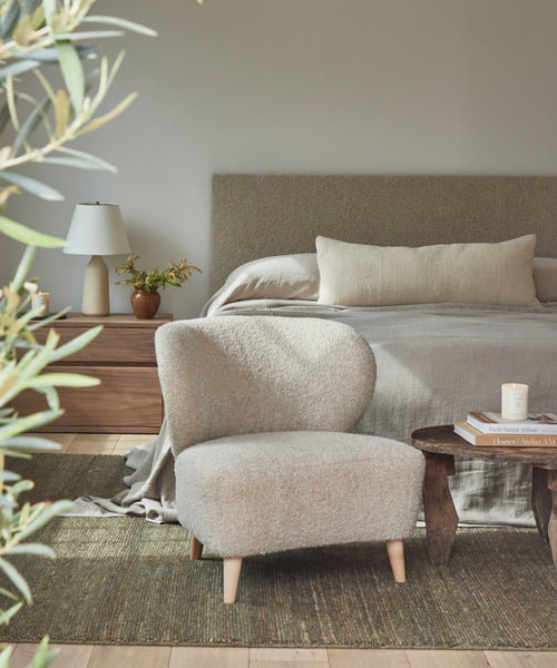 A cozy bedroom features a beige upholstered bed with neutral bedding, a Brentwood Chair in Belgian Wool Upholstery up front, and a wooden nightstand with a lamp and plant beside the bed. A small table with books sits in the foreground.