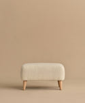 The Brentwood Ottoman is a compact, rectangular piece with off-white wool boucle upholstery and four short wooden legs, shown against a plain beige background.