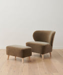 A modern brown upholstered chair and the Brentwood Ottoman, both featuring light wooden legs, are set on a light wood floor against a plain white wall.