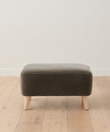 The Brentwood Ottoman, a compact rectangular piece in dark gray with light wooden legs, sits on a light wood floor by a white wall, adding stylish, modern upholstered accent seating to any space.