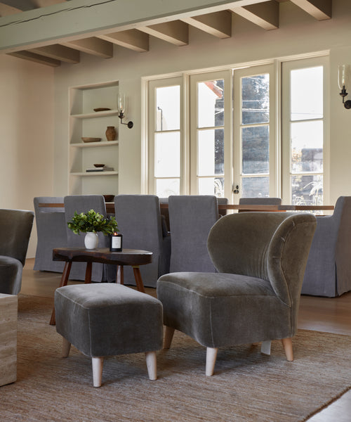 A cozy living room with a plush gray chair, Brentwood Ottoman, wooden side table topped with a plant and candle, neutral rug, dining table with gray slipcovered chairs, large windows, and open shelves in the background.