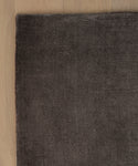 A corner of the dark brown Bristol Rug rests on a light wooden floor, showcasing the texture contrast between the soft rug and the smooth wood surface.