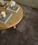 A round wooden coffee table with books, a lit candle, and a small vase of white roses sits atop the luxurious Bristol Rug, creating a cozy living room setting.