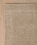 A corner of the Bristol Rug—a beige, textured, handwoven rug made in India—rests on a light wooden floor; only part of this silk rug is visible from above.