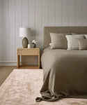 A modern bedroom features a beige bed, neutral pillows, light wood nightstand, ceramic lamp, and a small vase with white flowers atop the Bristol Rug, all set against white paneled walls.